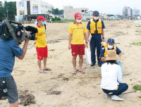 砂浜にいる人に声をかけているライフセーバーと監視員、それを撮影するテレビカメラ