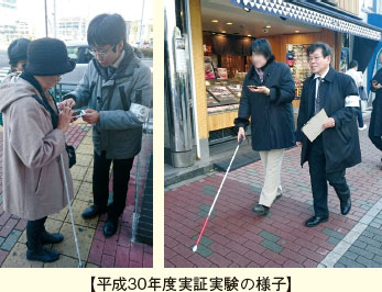 【平成30年度実証実験の様子】。スマートフォン端末及び白杖を持った被験者と付添人が歩道を歩いている様子の写真