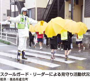 スクールガード・リーダーによる見守り活動状況。白い服を着たスクールガード・リーダーが横断歩道を横断中の児童に合図をしている写真
