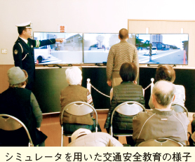 シミュレータを用いた交通安全教育の様子。交差点をイメージしたモニター画面に立つ人と指導員の写真