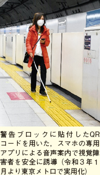 警告ブロックに貼付したQRコードを用いた，スマホの専用アプリによる音声案内で視覚障害者を安全に誘導（令和3年1月より東京メトロで実用化）。駅ホームでスマートフォンと白杖を持った人が警告ブロック上を歩いている様子