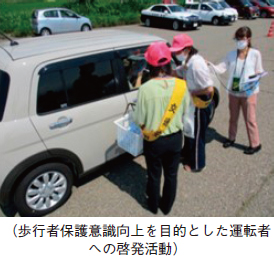（歩行者保護意識向上を目的とした運転者への啓発活動）。赤い帽子をかぶった参加者が自動車の運転手にノベルティを渡している