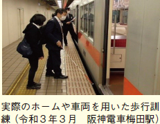 実際のホームや車両を用いた歩行訓練（令和3年3月　阪神電車梅田駅）。駅ホームで列車に乗ろうとしている白杖を持った人とそれを見守る人